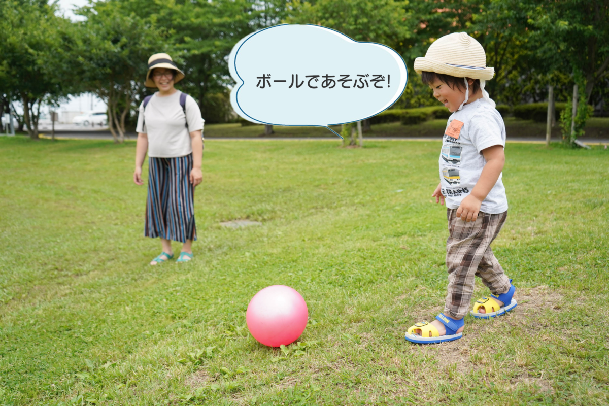ひだまり広場の芝生の上で、ボール遊びをする子どもと、近くで見守っているお母さんの写真。子どもが「ボールで遊ぶぞ！」と言っている。