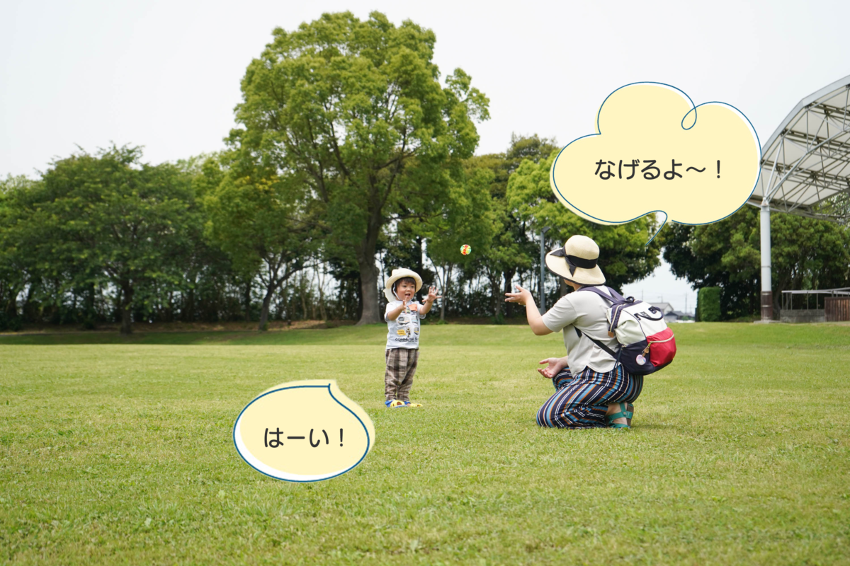 ひだまり広場の芝生の上で、キャッチボールをする子どもとお母さんの写真。お母さんが「投げるよ～！」と言い、子どもが「はーい！」と言っている。